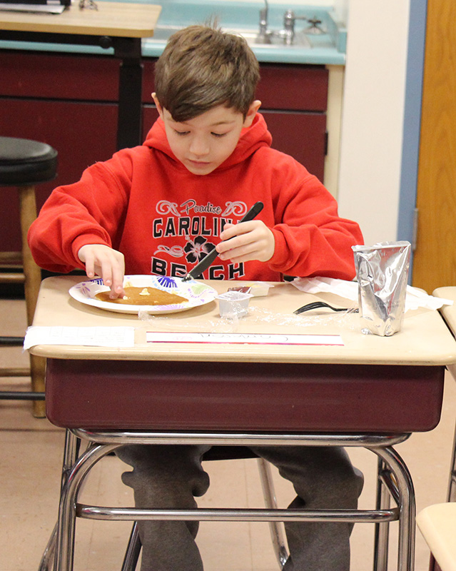 A fifth-grade boy with short dark hair wearing a red hooded sweatshirt sits at his desk eating a plate of pancakes.