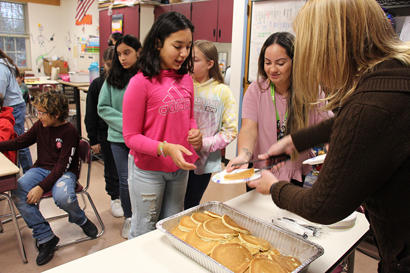 Fifth-grade girls stand in a line waiting to be served pancakes. The girl in front, with shoulder-length dark hair and a pink sweater, is reaching out to get a plate that is being handed to her.