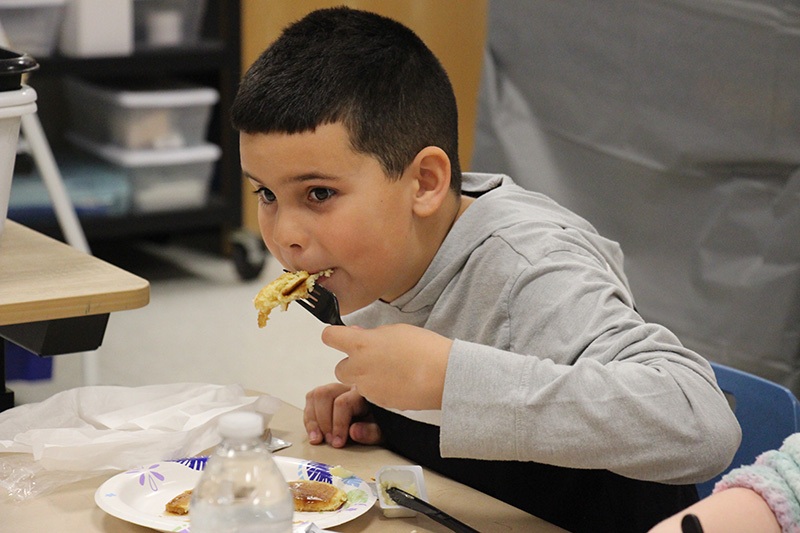 A second-grade boy with short dark hair and a gray long-sleeve shirt, puts pancakes into his mouth.