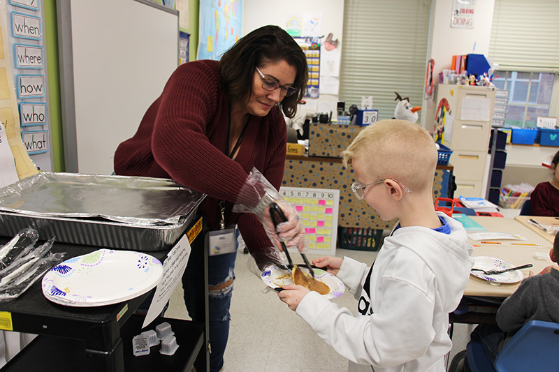 A woman wearing a maroon sweater, with shoulder-length dark hair, uses clippers to put a pancake on the plate of a boy with blonde hair, glasses and wearing a white hoodie.