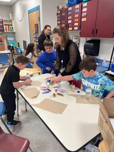 Elementary age kids work at a table together with a woman who is dressed in black. They are reaching for supplies on the table.