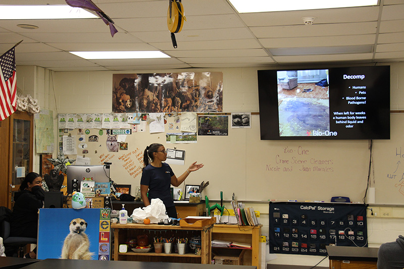 A woman dressed in dark clothing stands i front of a class of high school students. She is looking at a screen behind her that shows a crime scene.