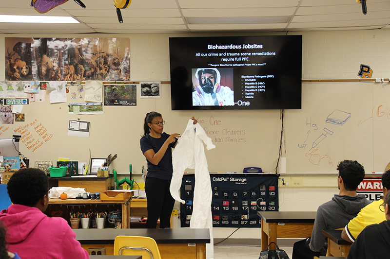 A woman dressed in dark clothing holds up a white hazmat suit. Behind her is a screen showing someone dressed in a hazmat suit. In front of her are high school students sitting at desks.