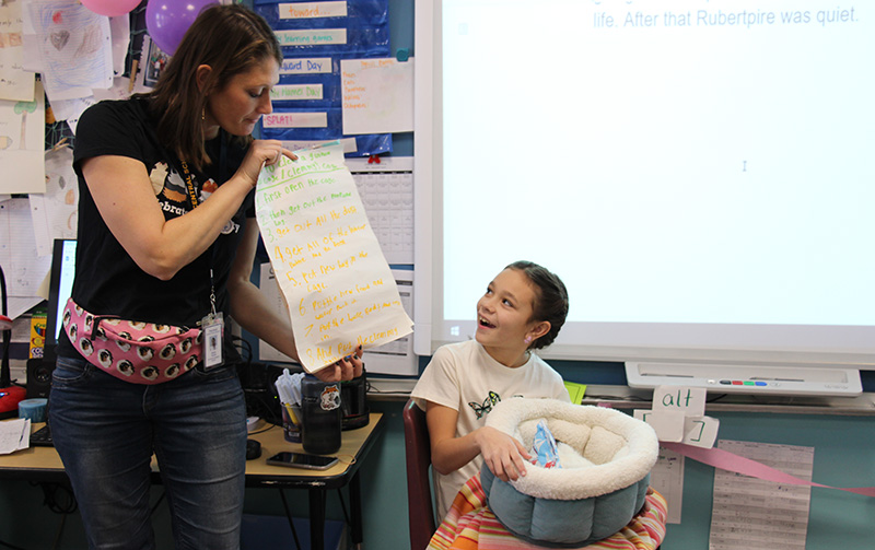 A girl smiles as she holds a plush carrier with a guinea pig in it. She turns and is reading from papers she created that are being held by her teacher, a woman with long dark hair.