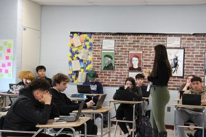 A woman with long dark hair, wearing a black shirt and green pants, talks to a group  of high school students, all seated at their desks.