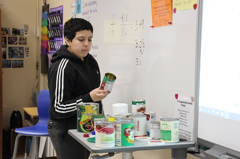 A boy wearing a black jacket with white stripes down the sleeves, looks at a small can he picked up from the many on a small table.