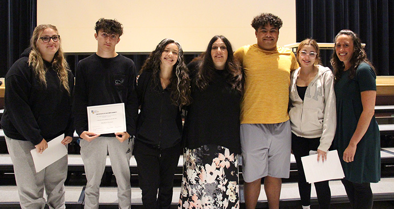 Five high school seniors stand with two teachers, all arm in arm.