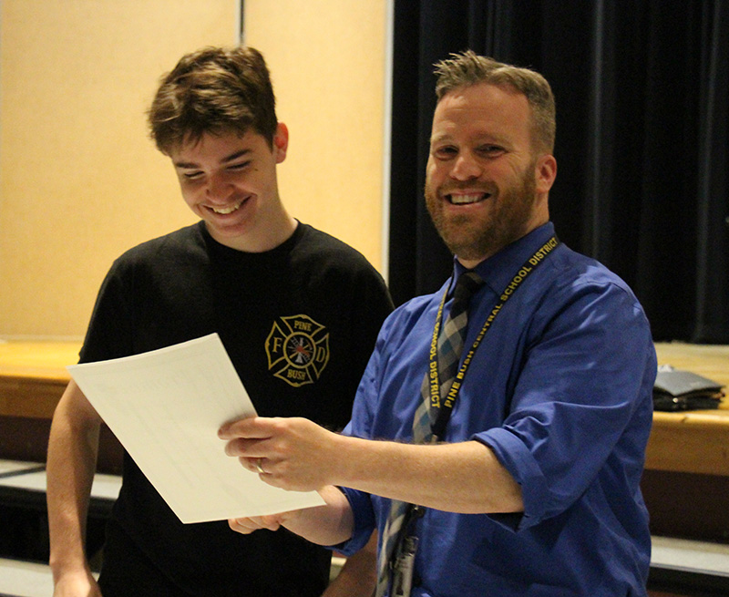 A high school senior young man smiles while looking at a piece of paper the man next to him is holding. The man is smiling.