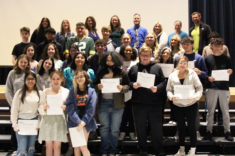 A group of about 35 mostly high school seniors and a few adults stand on a riser on a stage. Many of the students have certificates.