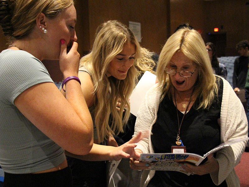 Two high school young women look at a yearbook with a woman. The woman is gasping and one of the young women has her hand over her mouth.