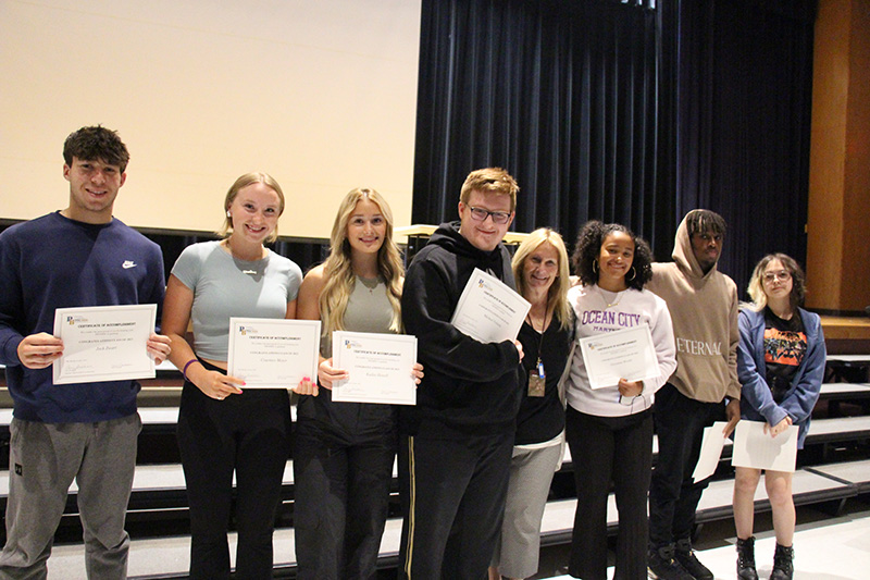 Seven high school seniors stand together arm in arm with one of their former teachers. The students have certificates.