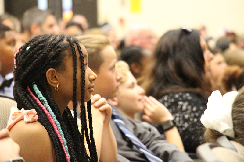 A girl with long braids sits listening to a speaker. Her hand is up to her chin. There are kids sitting in a row next to her.