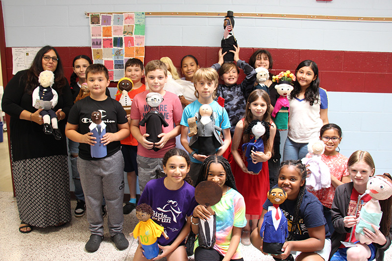 A large group of fifth-grade students holding their soda bottles decorated as famous people.