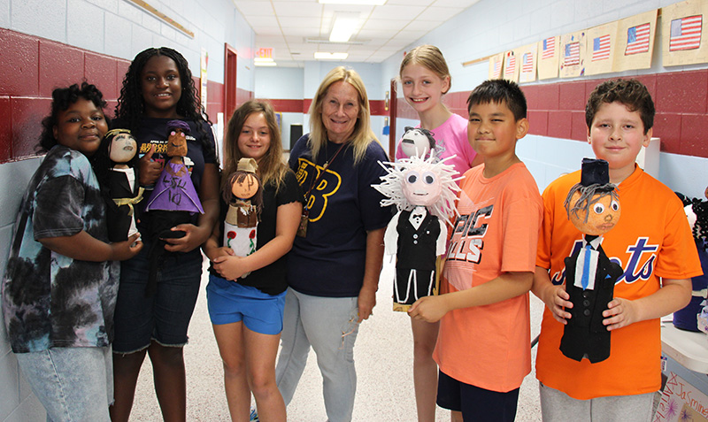 A group of six fifth-grade students hold their soda bottles decorated as famous people. They are standing with their teacher in the center.