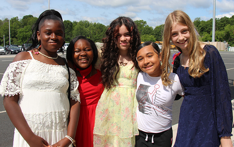 A group of five fifth-grade girls stand together, dressed nicely, and smile.