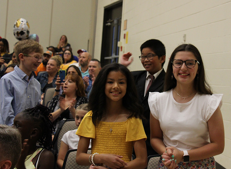 A group of four fifth-grade students stand among other students in an auditorium. Two girls in front smile.