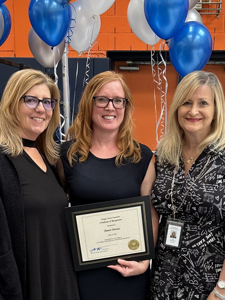 Three women stand together smiling. The woman on the left has shoulder'length blonde hair wearing a black shirt, the woman in the center has shoulder-length reddish hair and wearing a black dress, and the woman on the right has shoulder-length blonde hair and is wearing a black and white dress. The woman in the center is holding a certificate.