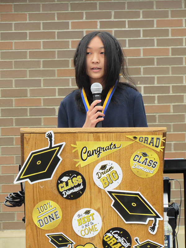 An eighth-grade girl with long dark hair holds a microphone and stands at a wooden podium with signs on it that say congratulations , grad and has graduation caps on it.