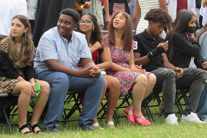 A group of six eighth-grade kids sit on folding chairs in the grass and watch to their right as others come to sit.