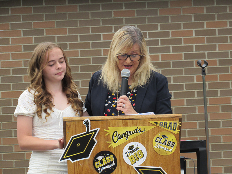 A woman with long blonde hair holds a microphone and looks down at a podium. Next to her is an eighth-grade girl with long blonde hair, wearing a white dress.