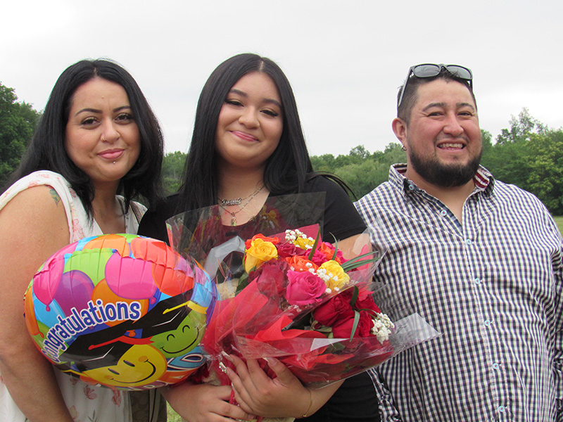 An eighth-grade girl with long dark hair holds a bunch of flowers and smiles. Next to her are a man on right and a woman on the left. They are smiling too.