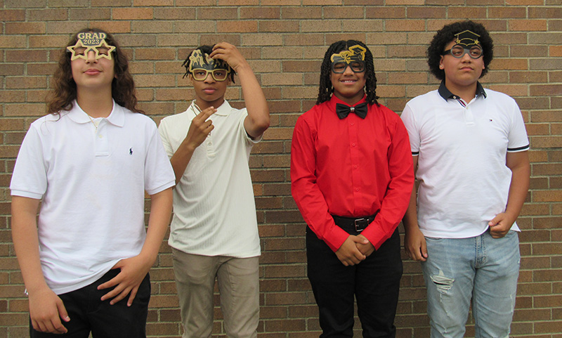 A group of four eighth-grade boys stand together and smile. They all have glasses on that say different things about graduation.