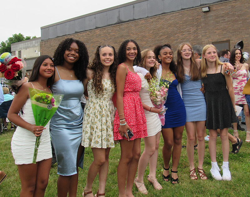 A group of eight girls, all dressed in dresses, have arms around each other. Many are holding flowers. They are all smiling.