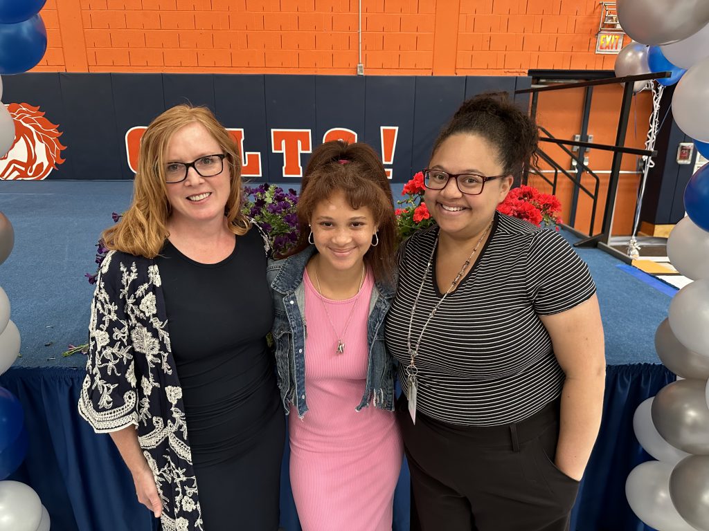 Two adults flank a middle school student in center. She is wearing a pink dress and her hair is pulled up. The woman are wearing black and white, glasses and all are smiling.