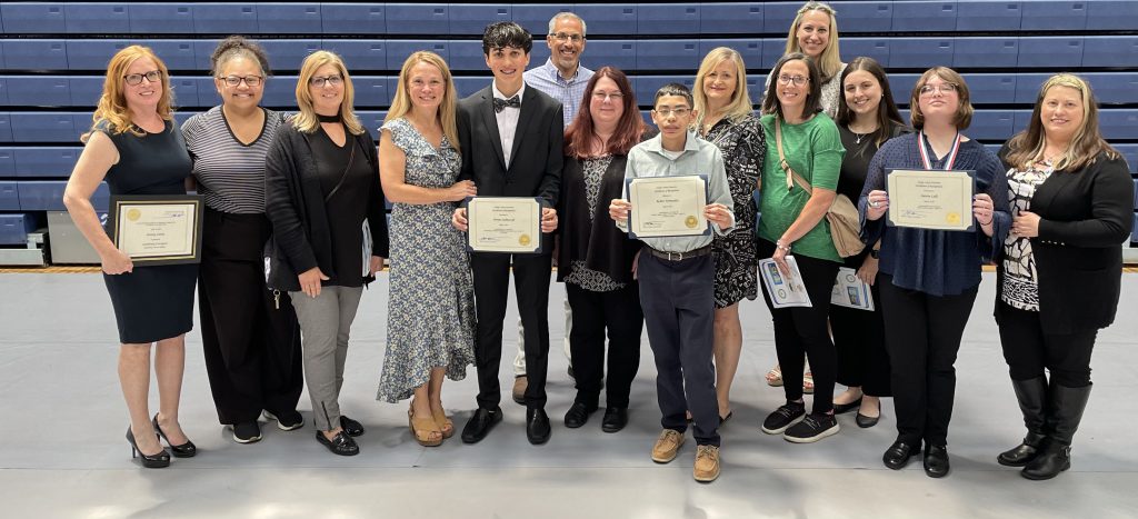 A group of 14 people stand together. There are  11 adults and three middle school students. The students are holding certificates as is the woman on the far left.