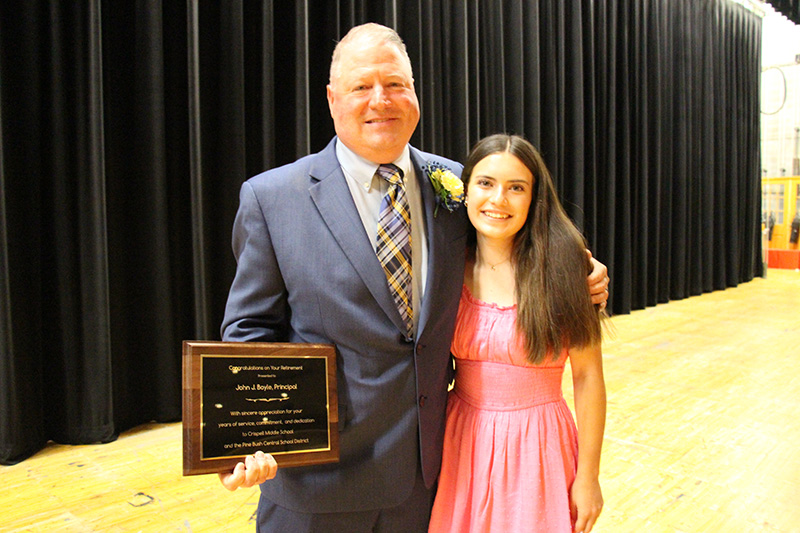 A man in a blue suit and tie holds a plaque. he ha his arm around the shoulder of an eighth-grade girl with long dark hair wearing a pink dress.