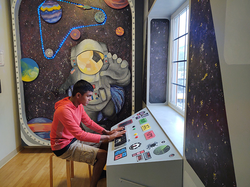 A middle school student sits at a control panel with the solar system painted in the background.