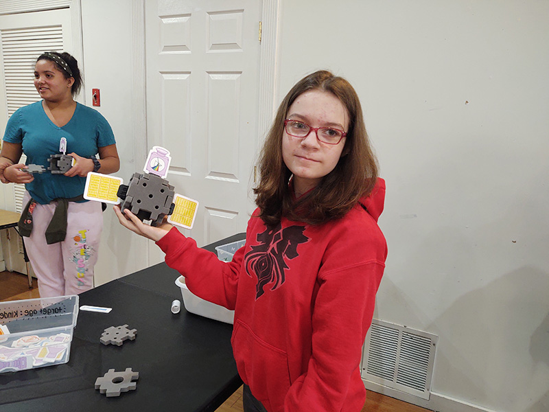 A middle school girl with long brown hair and glasses, wearing a red hoodie holds up a satellite she made.