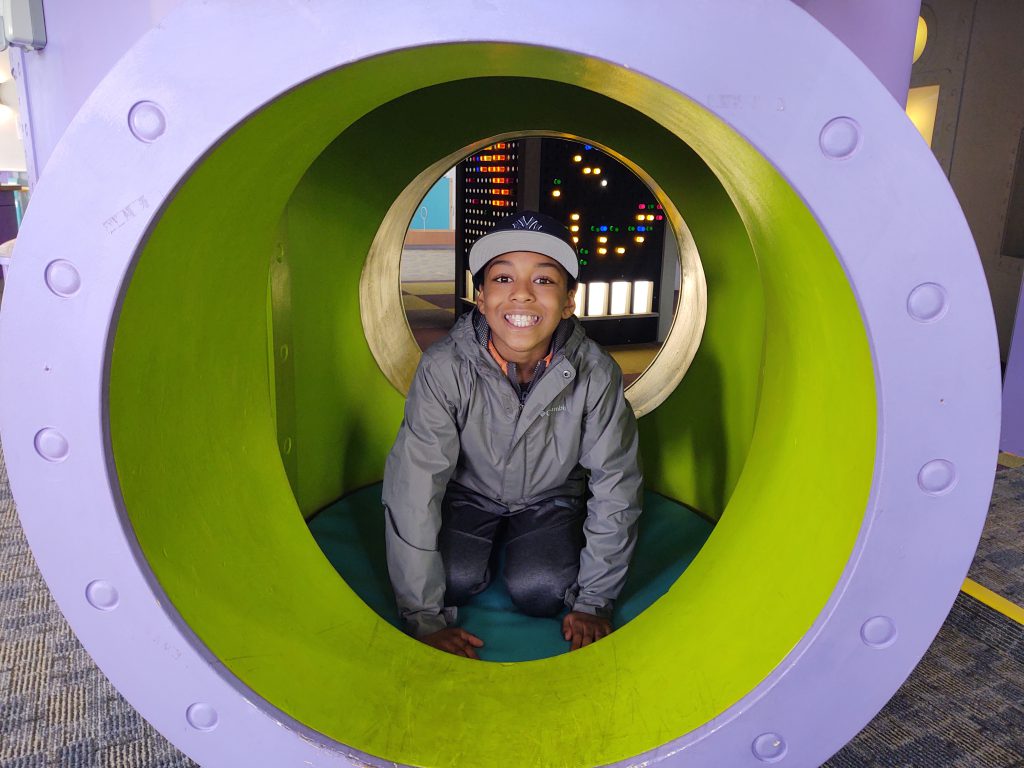 A middle school age boy, wearing a gray hoodie and a blue baseball cap smiles as he crawls through a large tube.