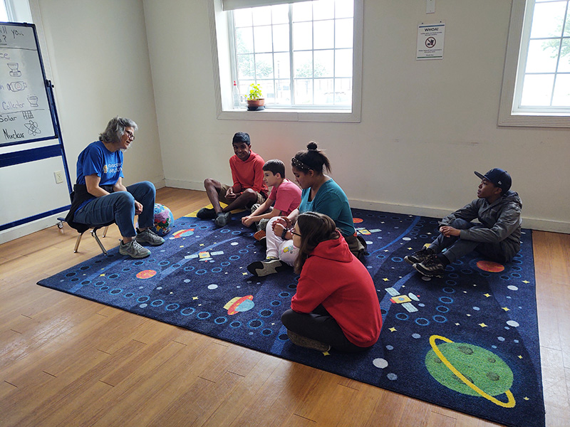 A group of four students and an adult sit on a solar system rug and listen to another adult teach.