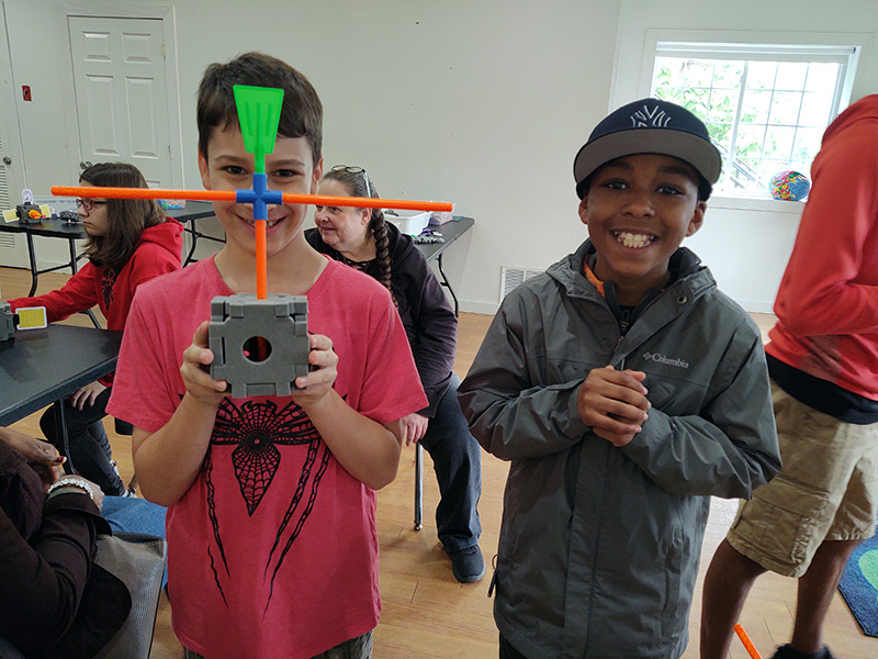 Two middle school boys smile for the camera. The boy on the left holds up a satellite he made while the boy on the right smiles.