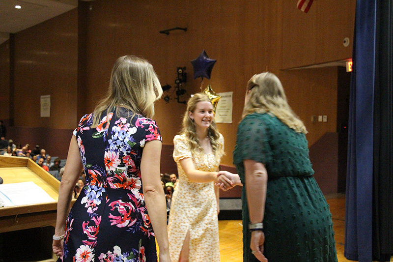 A young woman in a light colored dress shakes hands with a woman in a green dress as another woman in a flowered dress stands by.