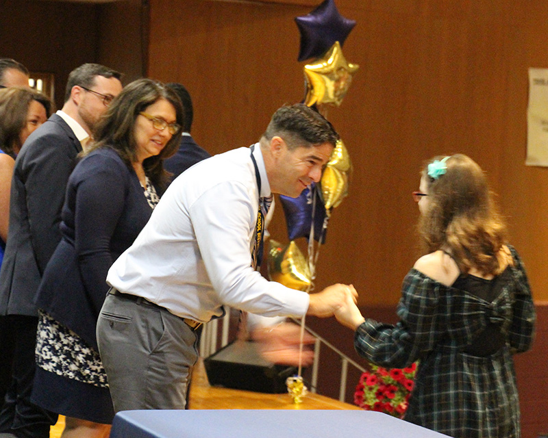 A man in a button-down shirt and tie bends over a bit and shakes hands with an eighth grade girl wearing a plaid dress.