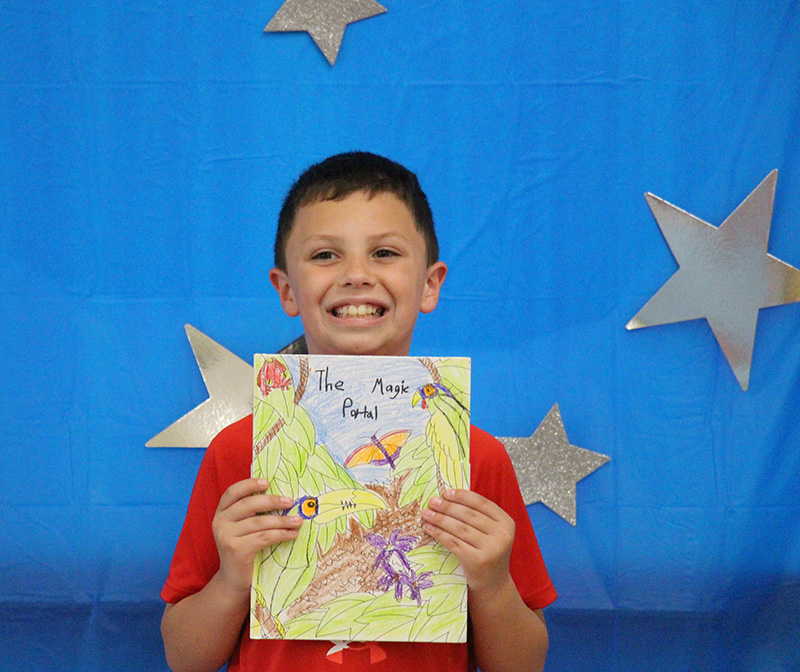 A third-grade boy with short dark hair wearing a red shirt holds up a book he wrote . The background is blue with silver stars on it.