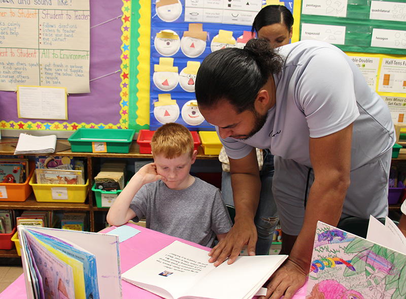 A third grade boy sits and shows a book he wrote and illustrated to a man who is leaning over the desk.