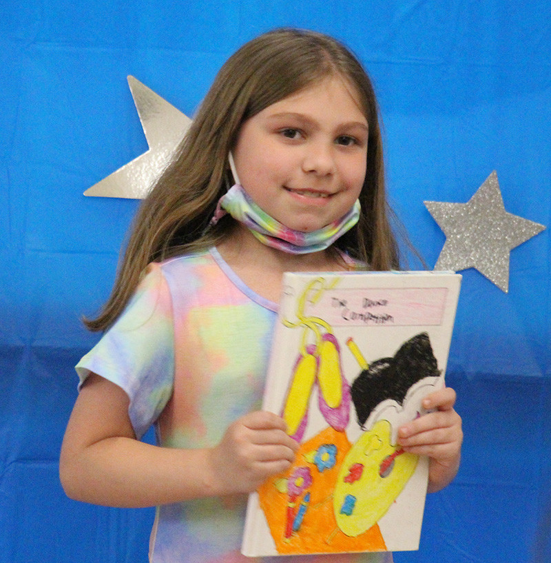 A third-grade girl wearing a tie-dye shirt smiles and holds up a book she wrote and illustrated.