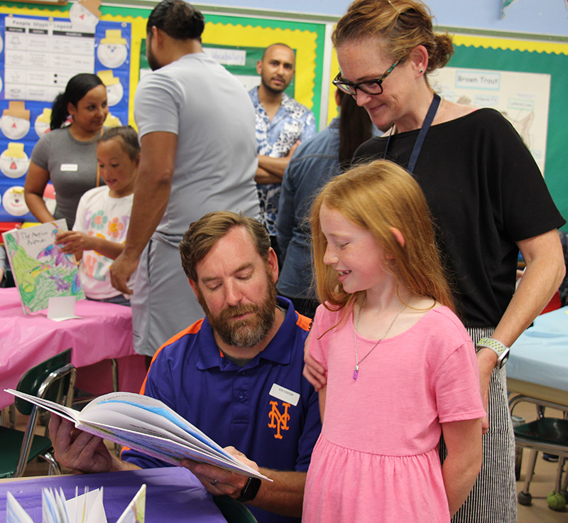 A third-grade girl with long red hair wearing a pink dress shows the book she wrote and illustrated to a man and woman.