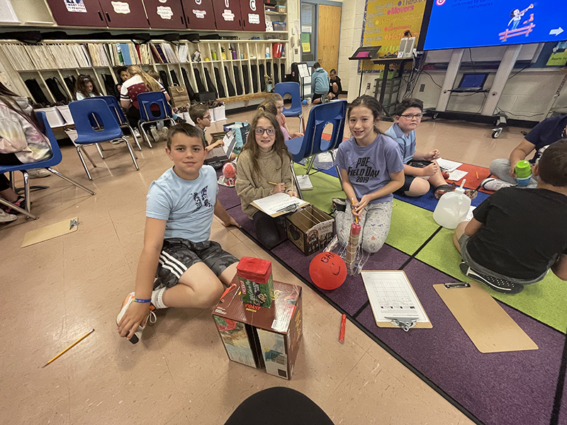 Three fourth grade students sit on the floor with recycled materials. They are smiling.