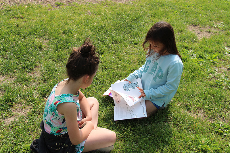 Two first grade girls sit in the grass as they read together.