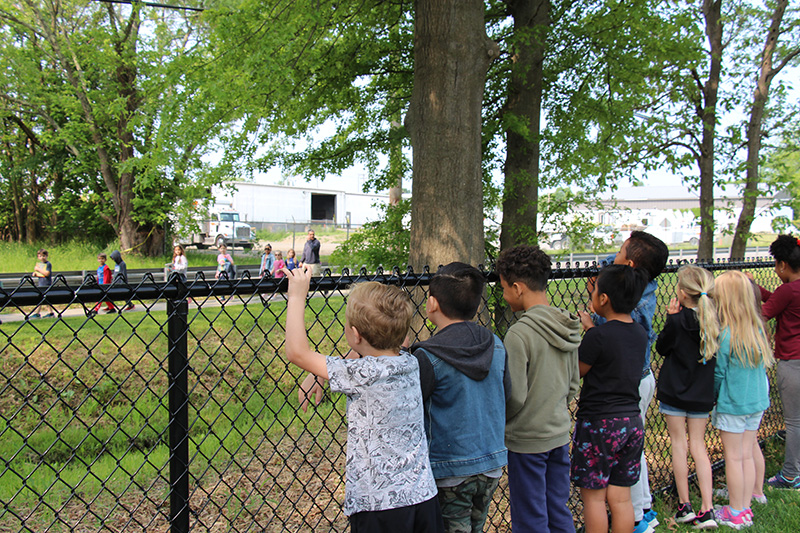 A group of first graders line a black fence looking at the students walking toward them.