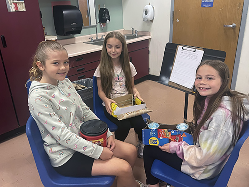 Three fourth grade students sit on chairs and hold recycled materials. They are smiling.