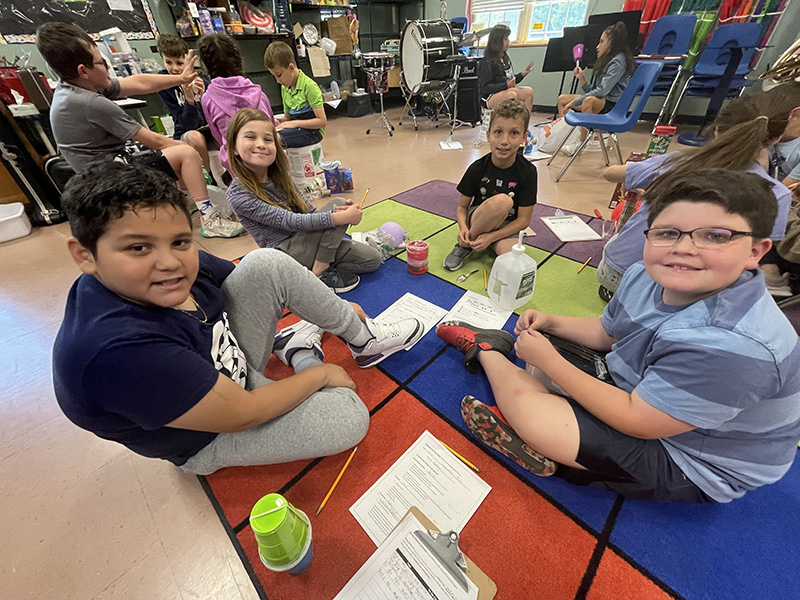 Four fourth grade students sit on the floor with recycled materials. They are smiling.