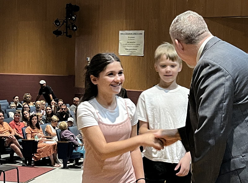 A fifth grade girl with dark hair, wearing a tan and white dress, shakes hands with a man with short hair and wearing a suit jacket.