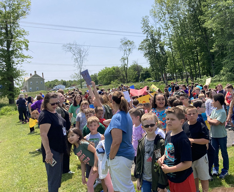 A large group of elementary students and staff line up waiting for the parade to pass by.