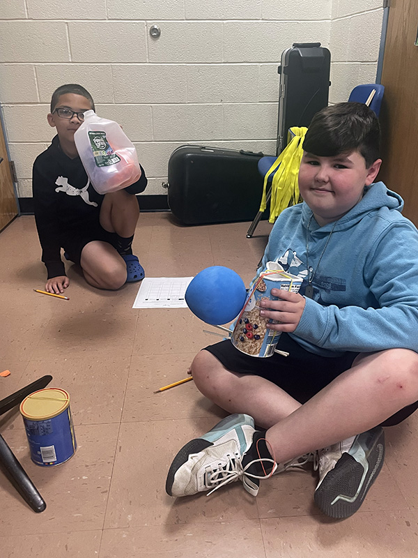 Two fourth grade students sit on the floor with recycled materials. They are smiling.