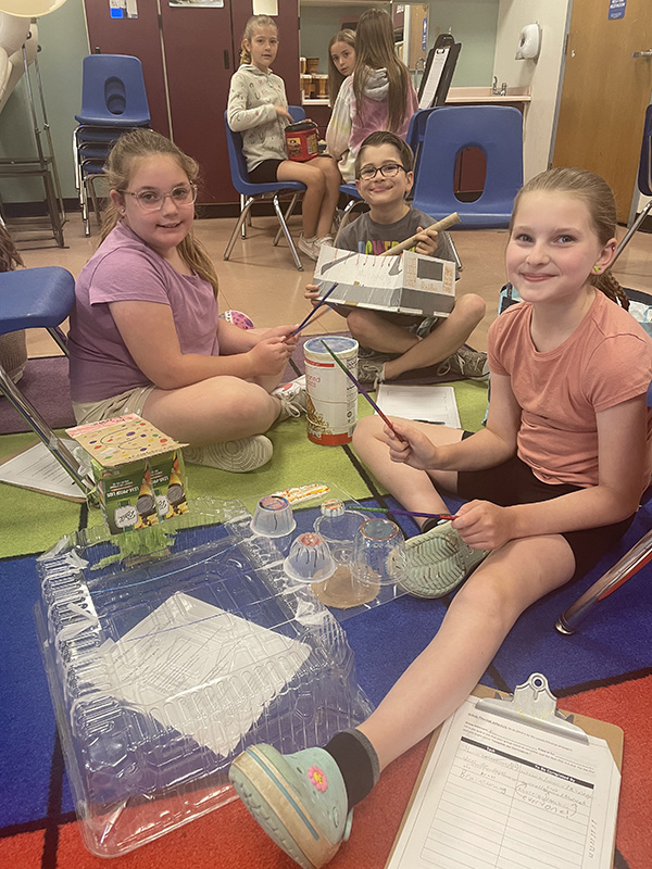 Three fourth grade students sit on the floor with recycled materials. They are smiling.